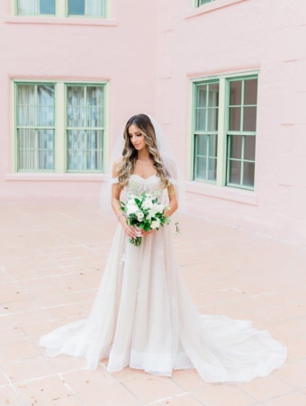 Elegant bride in white dress and veil poses in front of a pink building, exuding joy and beauty.